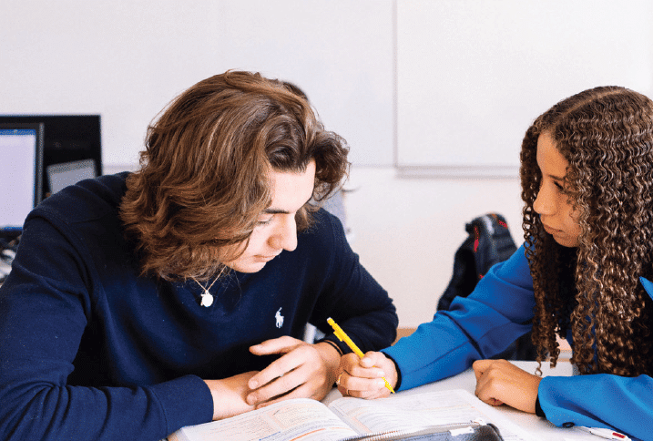 Deux élèves penchés sur un manuel ouvert, l'un prend des notes au crayon