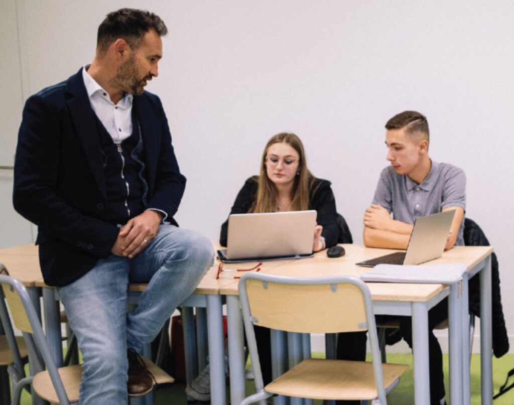 Un intervenant échange avec trois étudiants installés à leurs ordinateurs portables