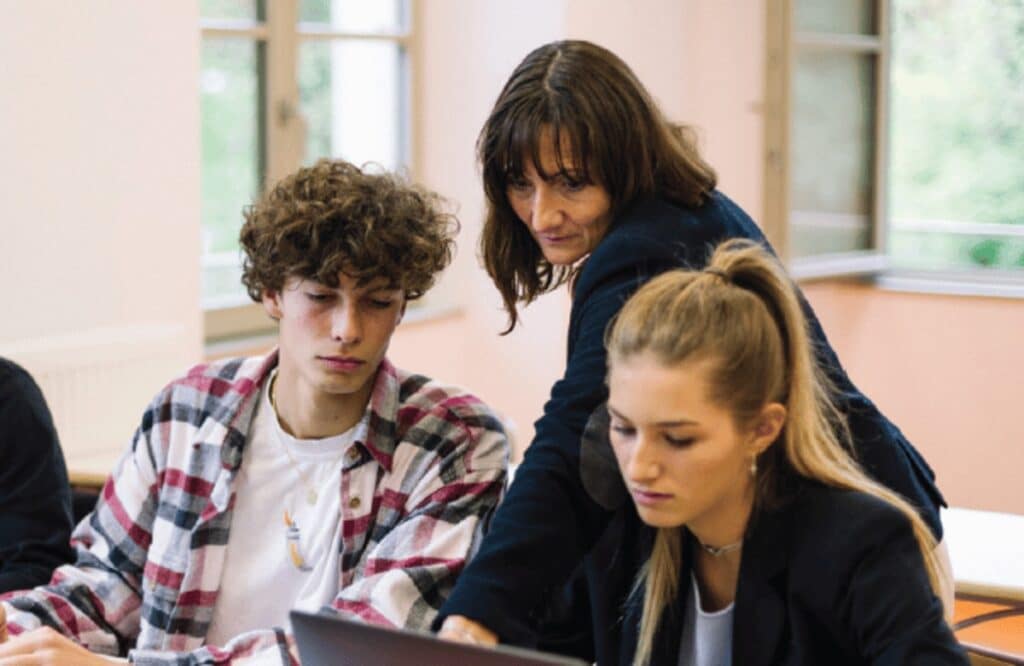 Une enseignante se penche vers deux élèves pour les aider devant leur ordinateur portable