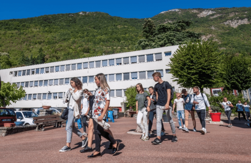 Des élèves traversent la cour devant le bâtiment du lycée, colline boisée en arrière-plan