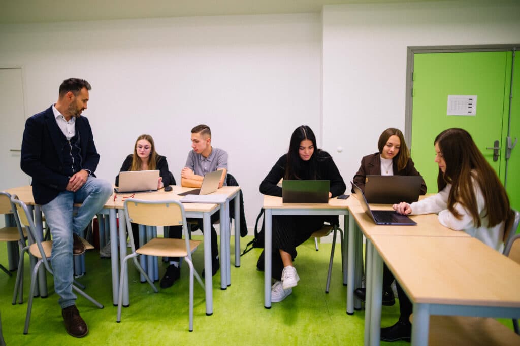 Salle de classe au sol vert, élèves installés par tables avec leurs ordinateurs portables